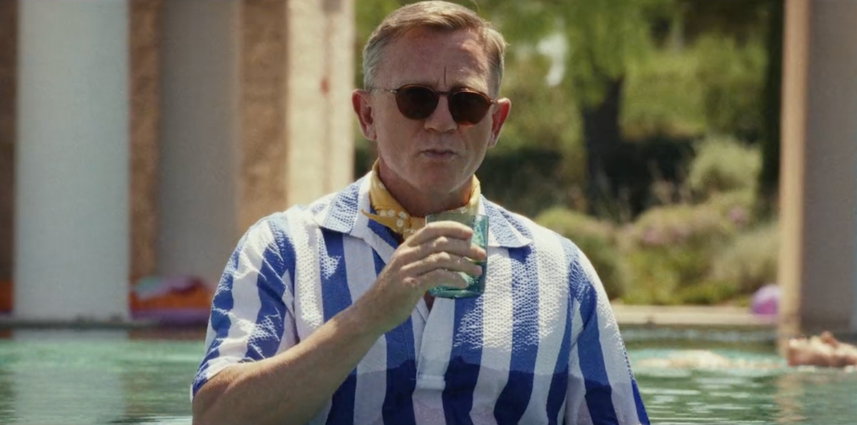 Image of Daniel Craig as Benoit Blanc in Glass Onion, standing in a pool in a striped bathing costume, holding a drink. 
