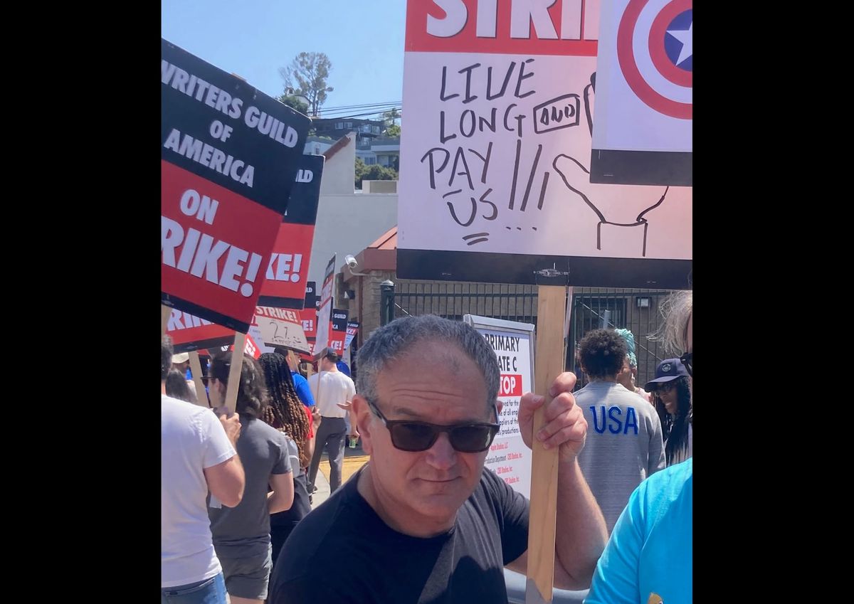 Photograph of Javi at the picket line, holding a sign that says "LIVE LONG AND PAY US" with a drawing of the Vulcan salute. Other strikers in the background holding signs.