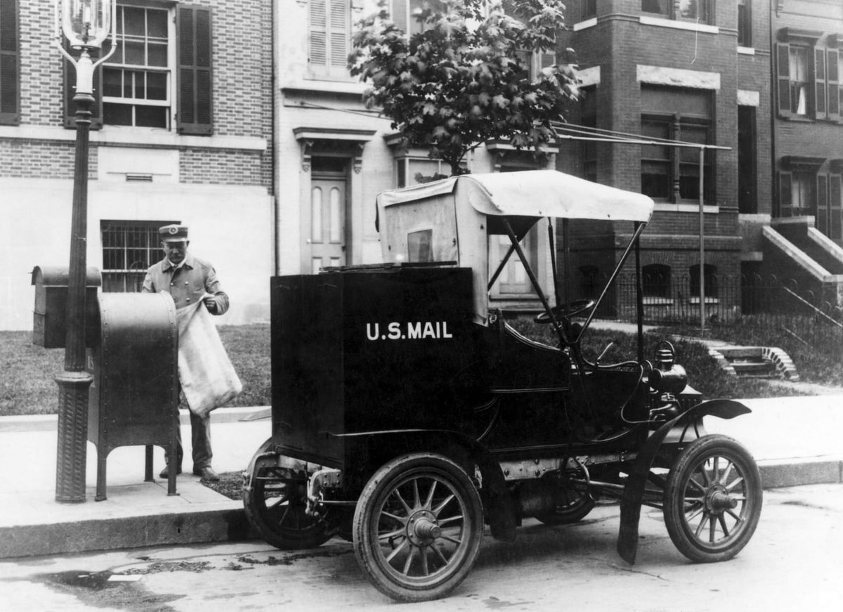 Black and white archival photo of a U.S. Mail truck and a mail carrier collecting from a curbside mailbox.