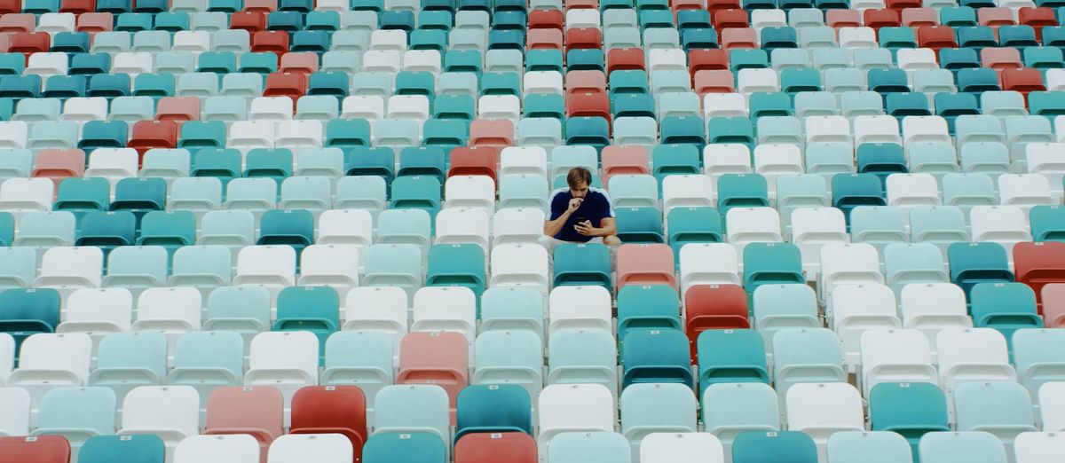 Photograph of a lone figure sitting in a section of a stadium with blue, white, and red chairs.