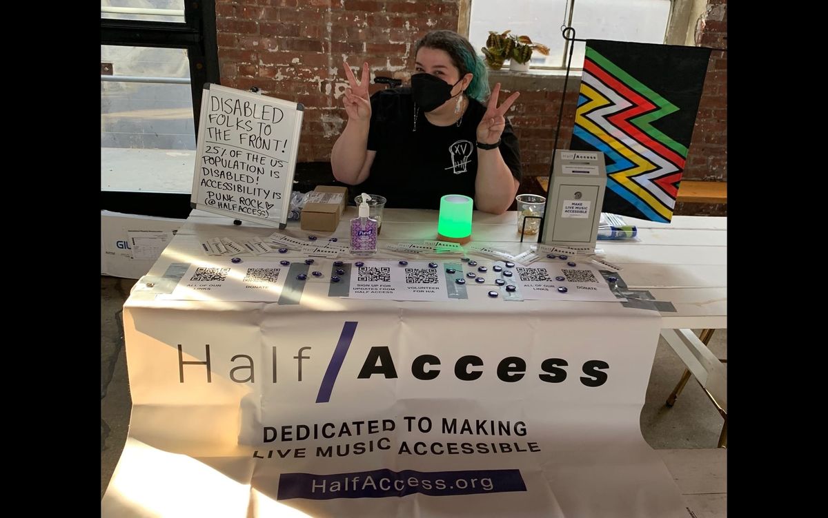 Photo of Valerie sitting at a table making peace signs with her hands. Banner on table reads: Half/Access Dedicated to Making Live Music Accessible HalfAccess.org.