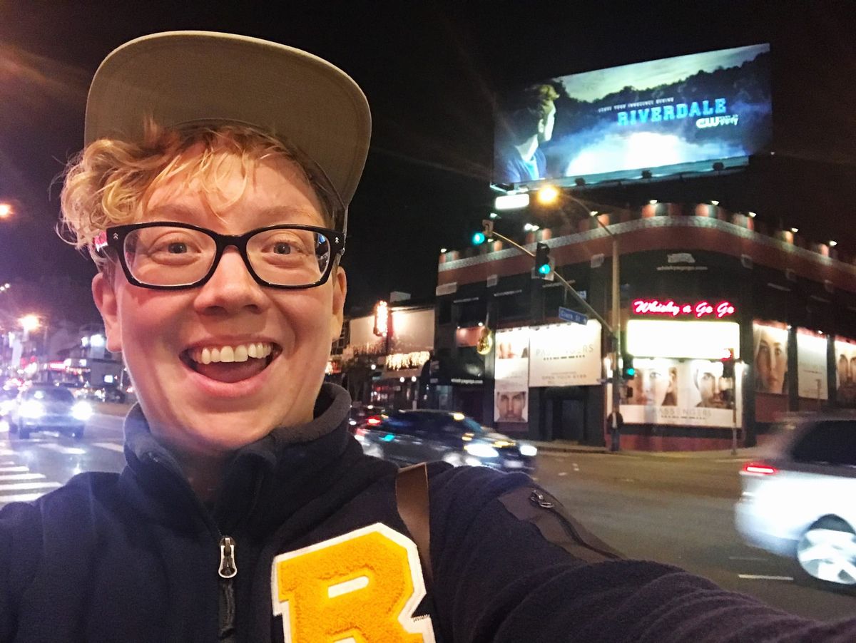 Britta Lundin, wearing a Riverdale letter jacket, grinning in front of a Riverdale billboard. 