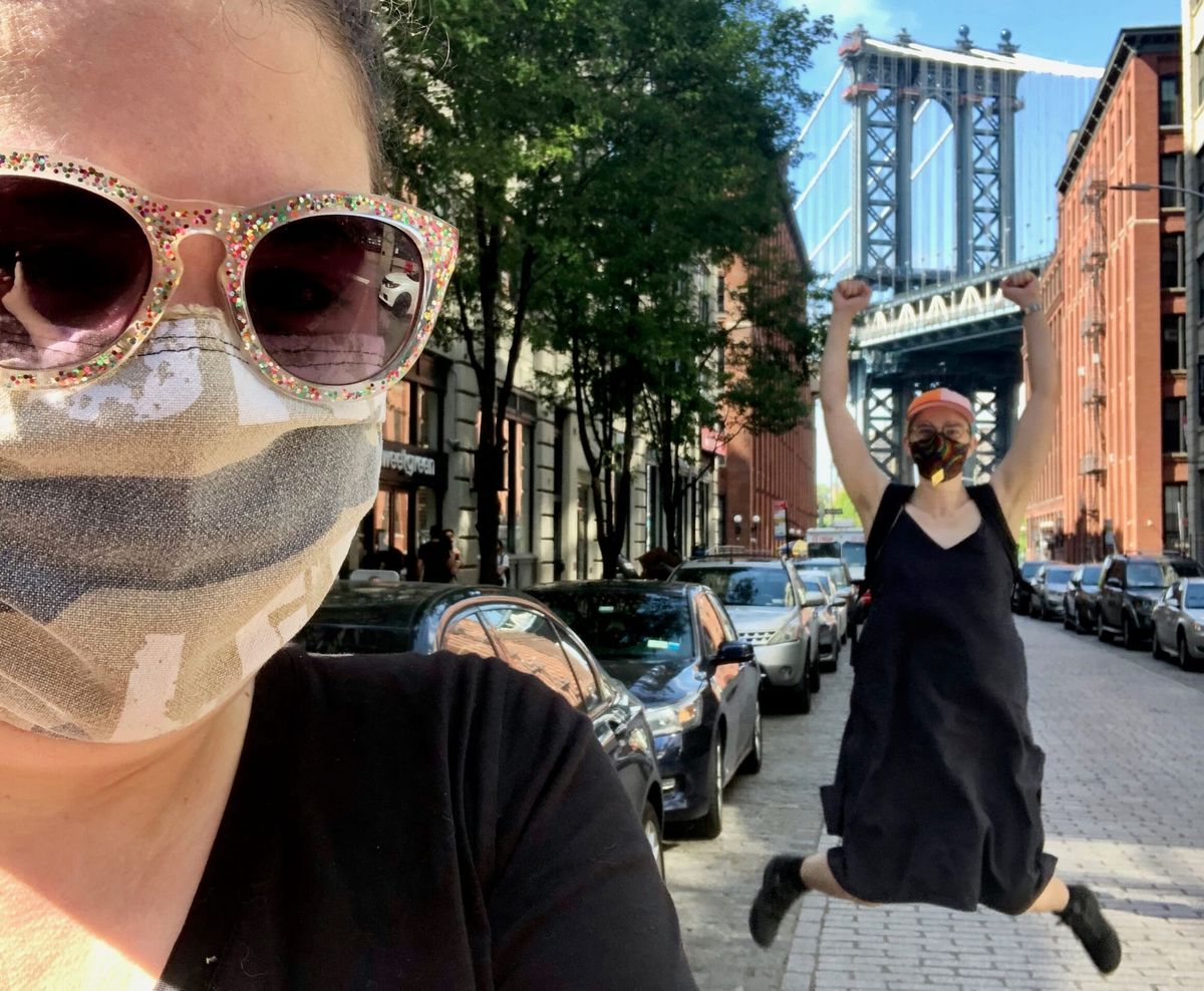 Selfie taken by Elizabeth in the foreground, with Flourish in the background jumping in the air, backdropped by the Manhattan bridge. Both wear masks and Elizabeth wears glittery sunglasses.