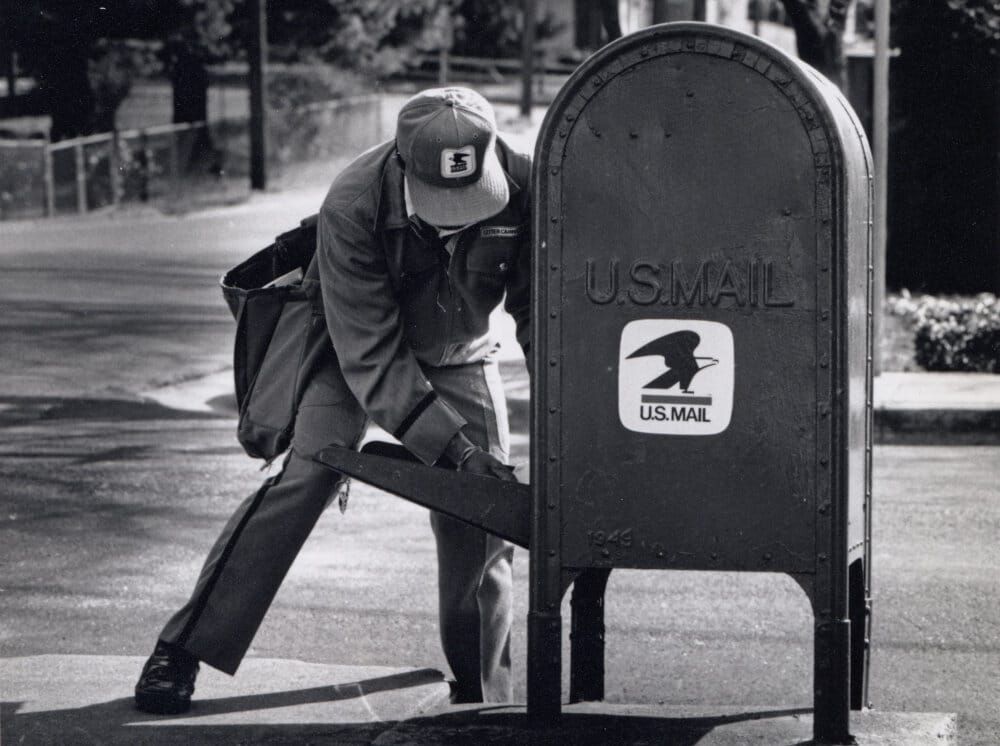 Black and white image of a USPS postal worker getting mail from a box. 