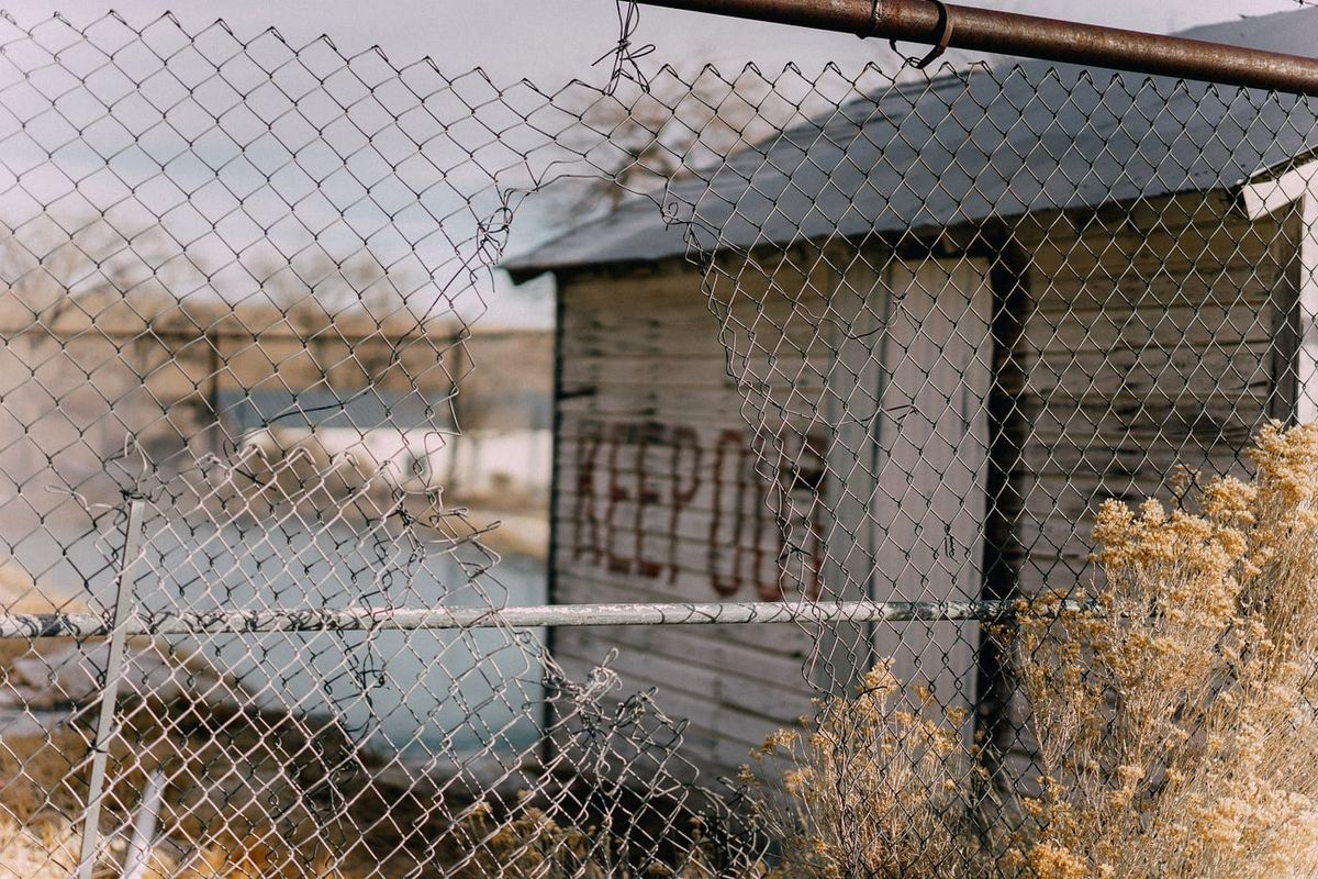 Photograph of a derelict building with a torn fence. Spray painted on the building wall is KEEP OUT. 