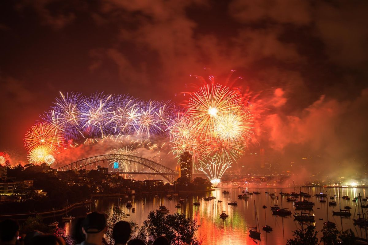 Image of fireworks going off over a harbor, lighting up the sky in shades of orange, red, and purple.