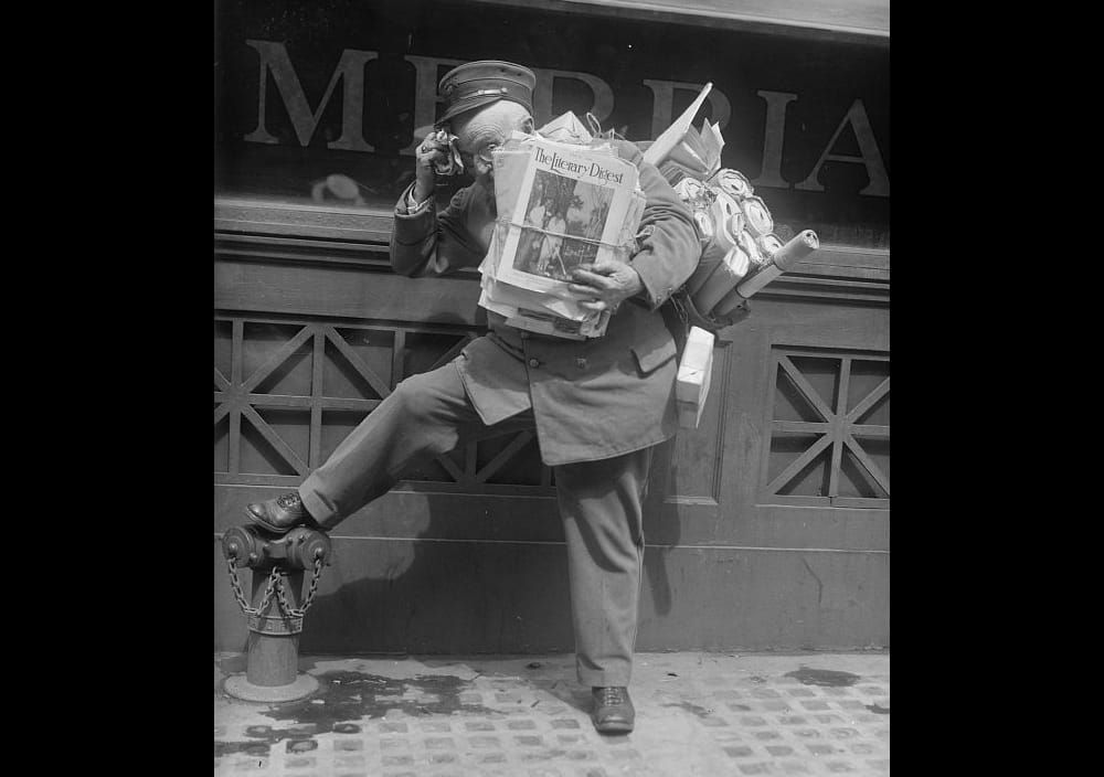 Black and white archival image of a postal carrier with his arms full of letters and magazines, his foot resting on a fire hydrant.