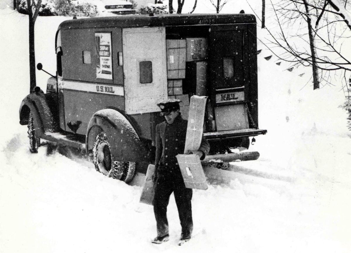 Black and white archival photo of a mail carrier delivering packages through the snow. 