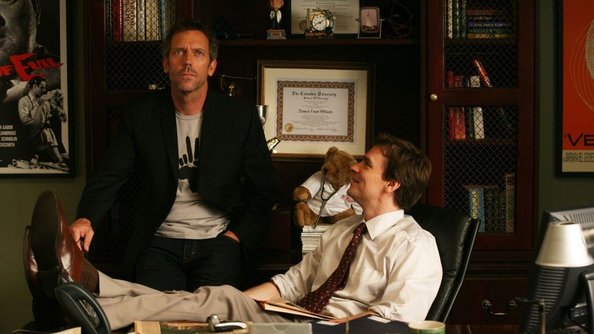 Promotional photograph for House, MD featuring Wilson seated at his office desk, feet up, looking at House, seated on his bookshelves and looking upwards.