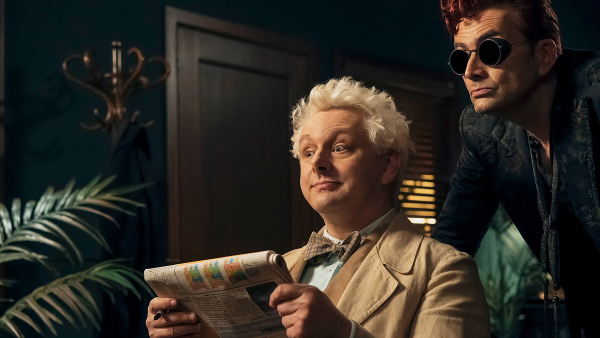 Production still of Aziraphale and Crowley looking at someone offscreen. Aziraphale holds a newspaper.