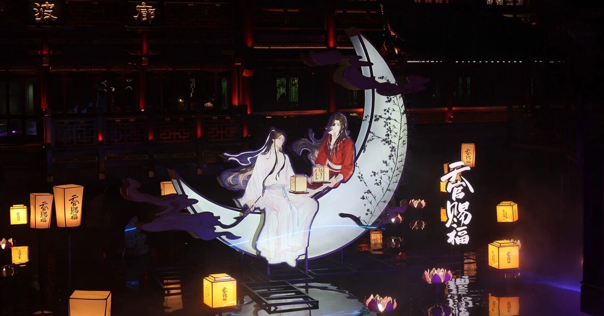 A nighttime photo of a display of the main characters from Heaven Official's Blessing. The display is a 3-D stand of the couple sitting together on the crescent moon together.