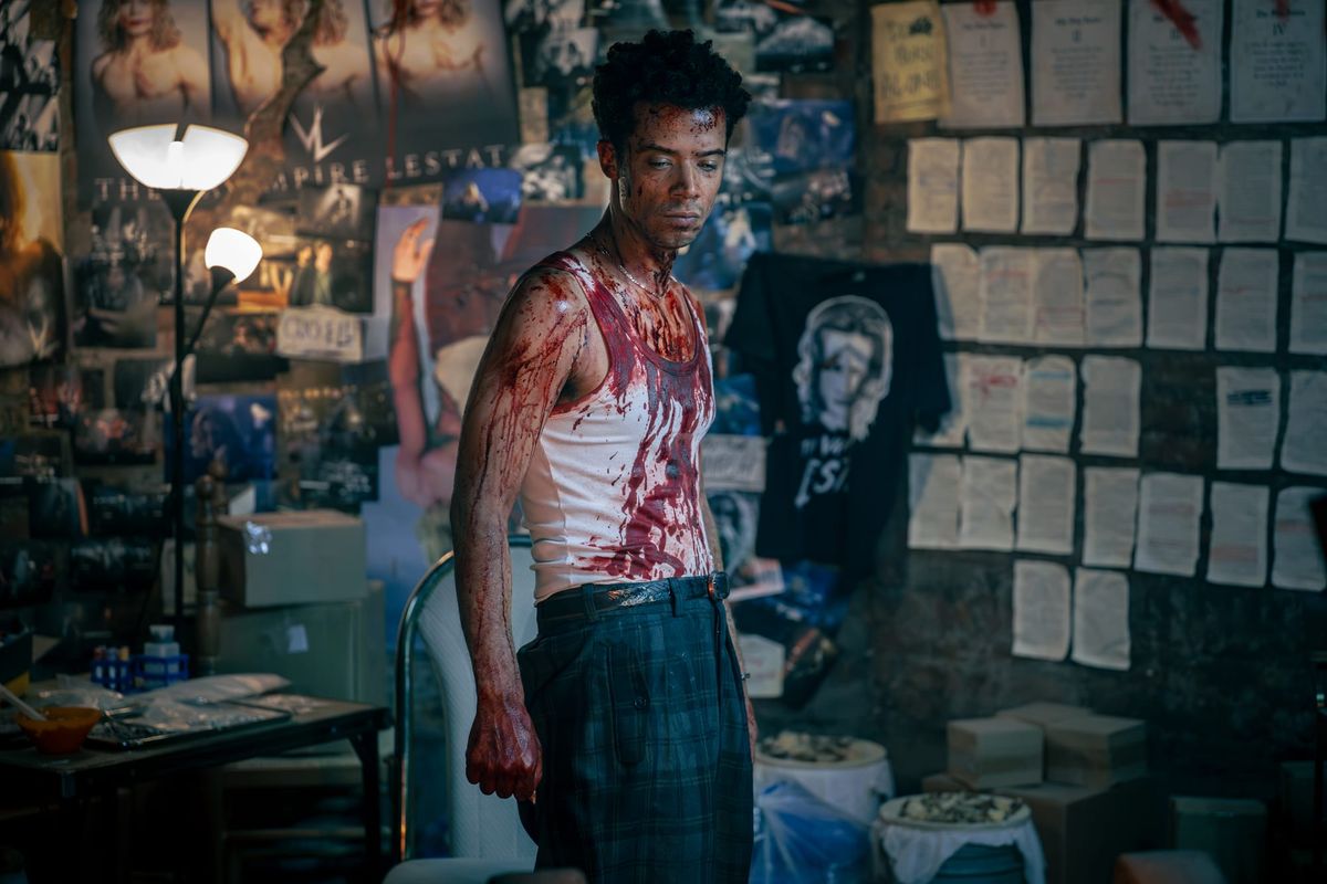Production still featuring Louis in a white shirt and dark plaid pants, covered in blood, with a wide range of Lestat merch and print-outs on the walls.
