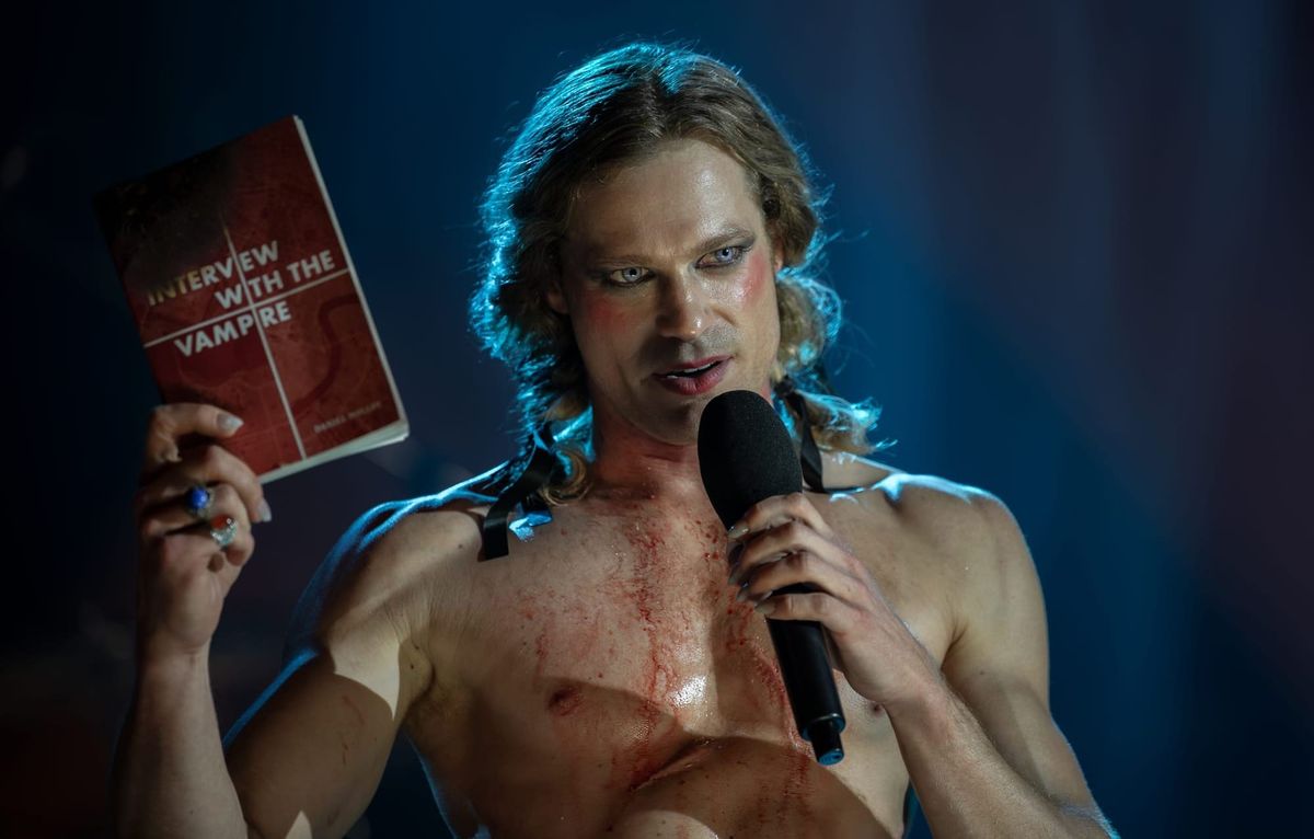 Image of Lesat onstage, shirtless with his hair in pigtails, holding a mic in one hand and the IWTV book in the other.