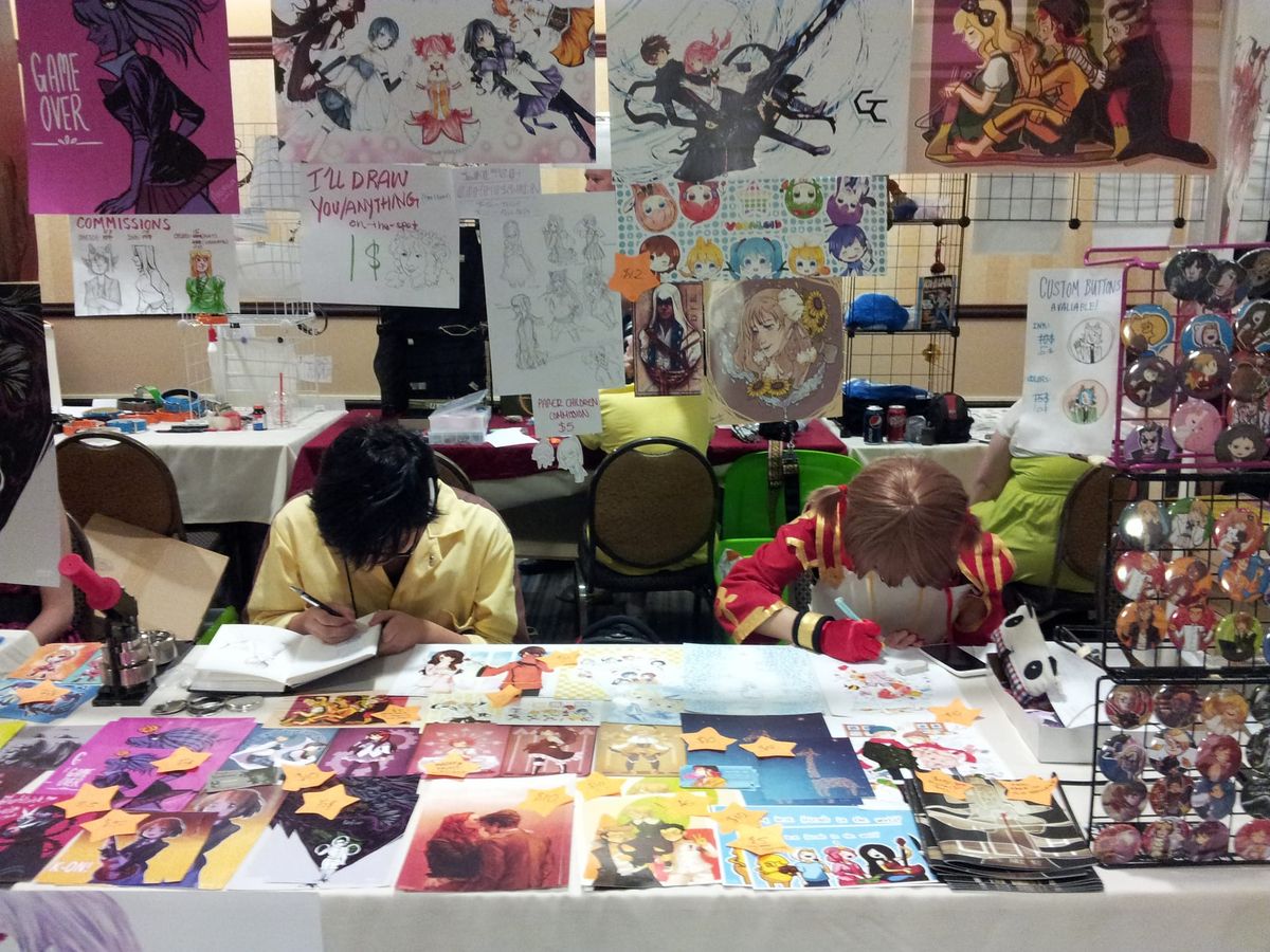 Two young artists bent over their sketchbooks while working at a booth at Animinneapolis 2013. The booth is colorful and full of prints and pins of anime characters.