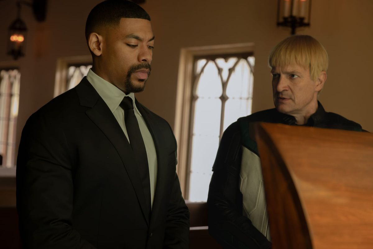 Production still of John and Guy dressed in black, standing in front of Hal's open coffin at the funeral.