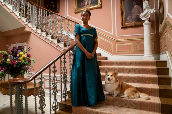 Promotional still from Bridgerton featuring Kate in a blue Empire-waist gown standing at the base of a set of carpeted stairs, a corgi sitting on one of the stairs near her feet.