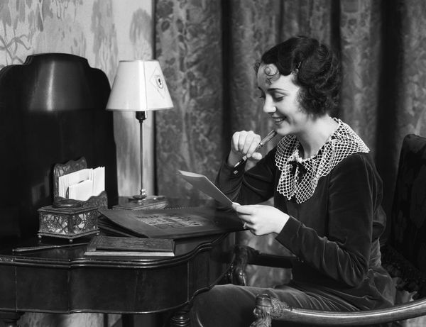 Black and white image from the 1930s of a woman sitting at a desk holding a paper and pen, smiling.