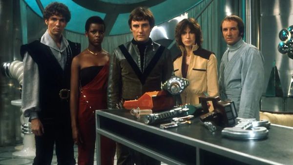 Promotional photo of the cast, standing behind a silvery table with props.. See the caption for actors' names.