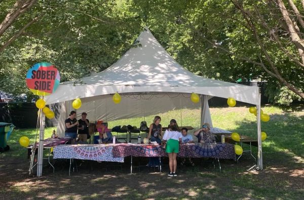 Photograph of a tent labeled SOBER SIDE with several people sitting at tables draped in colorful cloth, with yellow balloons on the edges of the tent.