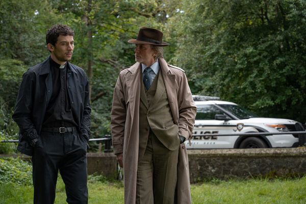 Still from Wake Up Dead Man featuring Father Jud and Benoit Blanc walking together on the church grounds, a police car in the background.