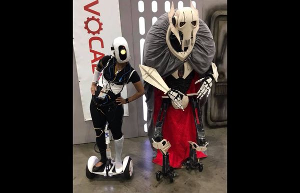Photograph of two figures cosplaying at a con. One (Joan) stands on a Segway. The other has an elaborate headpiece and sword.