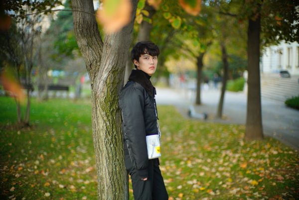 Photograph of Liang standing with their back to a tree, with autumn leaves on the ground.