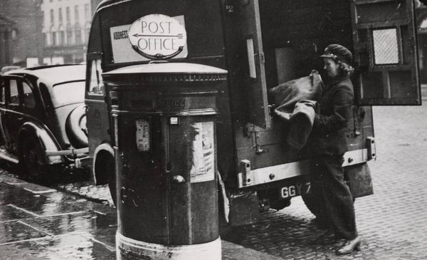 Black and white archival image of a female postal worker lifting a bag from a mail truck.