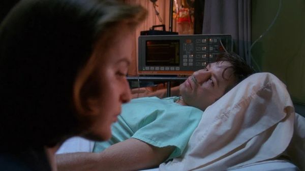 Screengrab from the X-Files. Scully in the foreground, blurred; Mulder in focus behind her in a hospital bed.