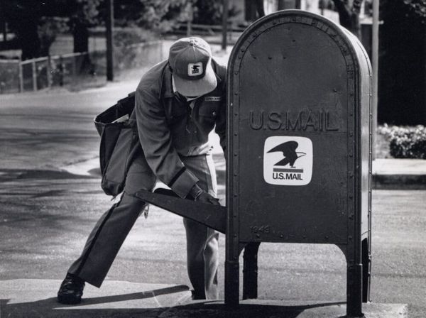 Black and white image of a USPS postal worker getting mail from a box. 