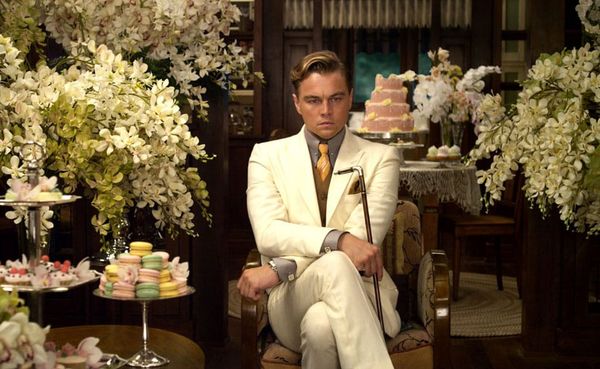 Image of Leonardo DiCaprio as Jay Gatsby sitting cross-legged in a white suit, looking annoyed, while surrounded by many flowers and trays of cakes.