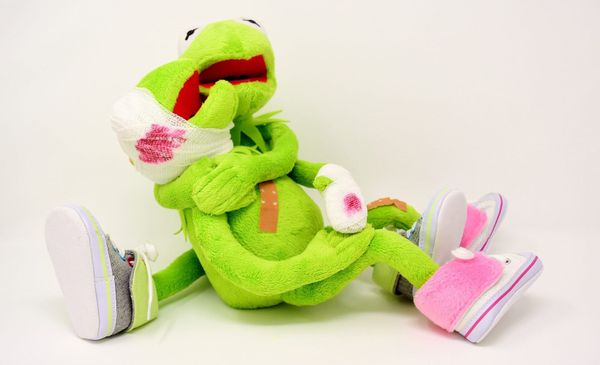Image of two stuffed Kermits, one with a bloody bandaged head and hand laying in the arms of the other. Both look distraught.