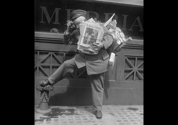 Black and white archival image of a postal carrier with his arms full of letters and magazines, his foot resting on a fire hydrant.