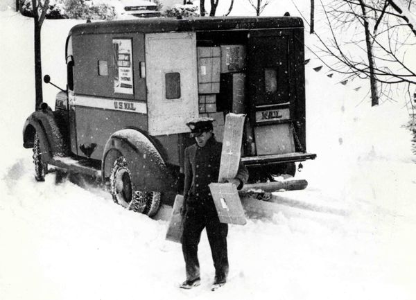 Black and white archival photo of a mail carrier delivering packages through the snow. 