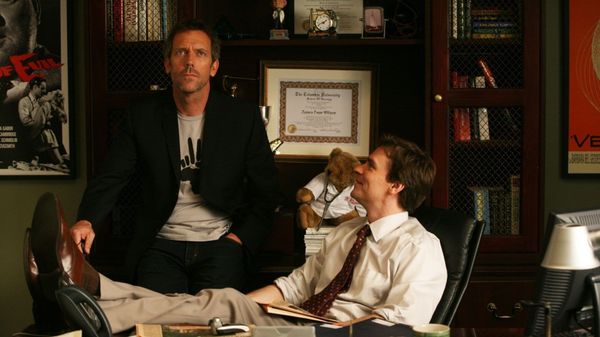 Promotional photograph for House, MD featuring Wilson seated at his office desk, feet up, looking at House, seated on his bookshelves and looking upwards.