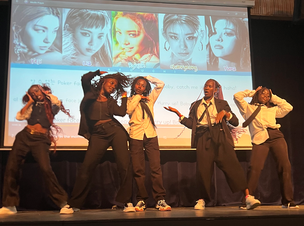 K-pop fans onstage at K-pop Fest perform to the music of K-pop band Itzy, whose images are shown on an overhead screen behind the performers.