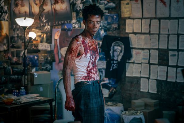 Production still featuring Louis in a white shirt and dark plaid pants, covered in blood, with a wide range of Lestat merch and print-outs on the walls.