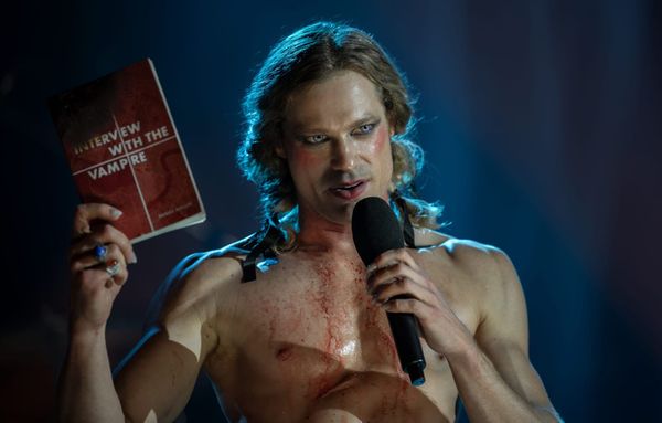 Image of Lesat onstage, shirtless with his hair in pigtails, holding a mic in one hand and the IWTV book in the other.