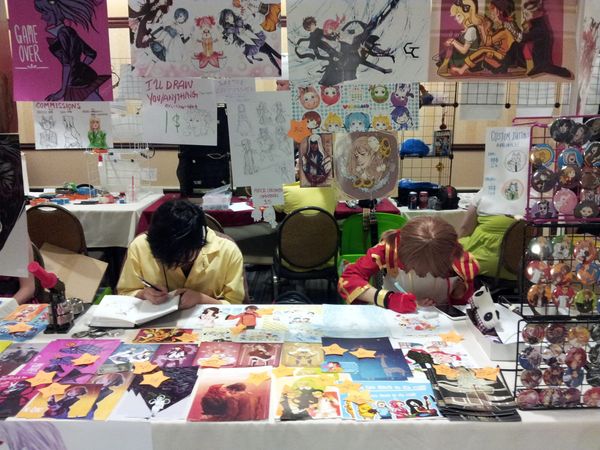 Two young artists bent over their sketchbooks while working at a booth at Animinneapolis 2013. The booth is colorful and full of prints and pins of anime characters.