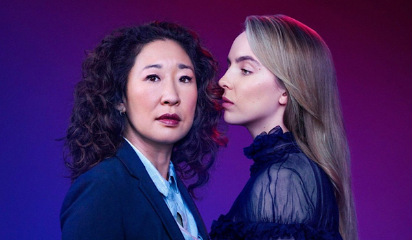 Jodie Comer stares at Sandra Oh, who is looking away from her, off to the left of the camera. The background is blue to purple ombre and they are dressed masc and femme by contrast.