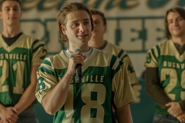 Production still of Noah in his gold and green Rushville football jersey, holding a mic. Three teammates stand in the background in matching jerseys.