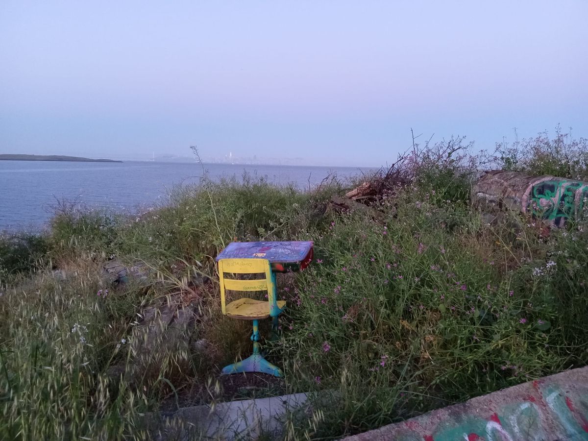A yellow spray-painted chair is placed in front of a decaying desk amidst tall grass, overlooking the Bay, with the San Francisco skyline faintly visible in the background.