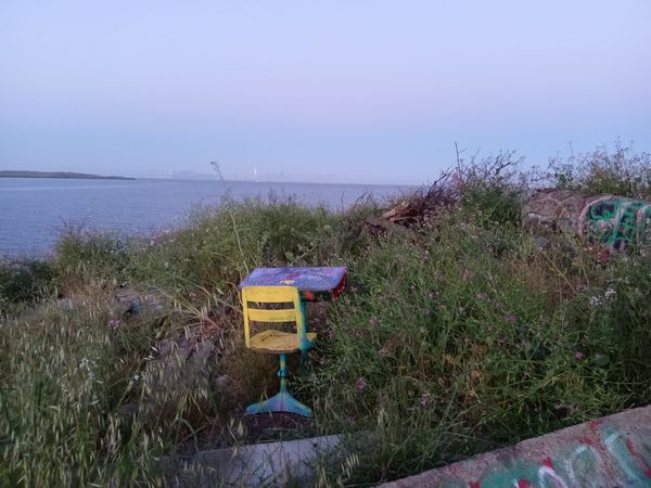 A yellow spray-painted chair is placed in front of a decaying desk amidst tall grass, overlooking the Bay, with the San Francisco skyline faintly visible in the background.