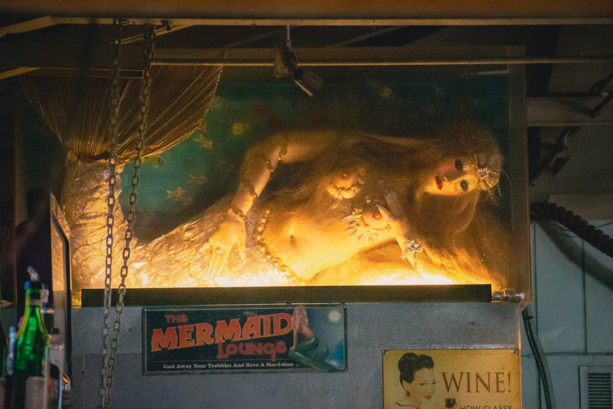 A photo of an illuminated mermaid mannequin in a glass fish tank as decor in a bar room.
