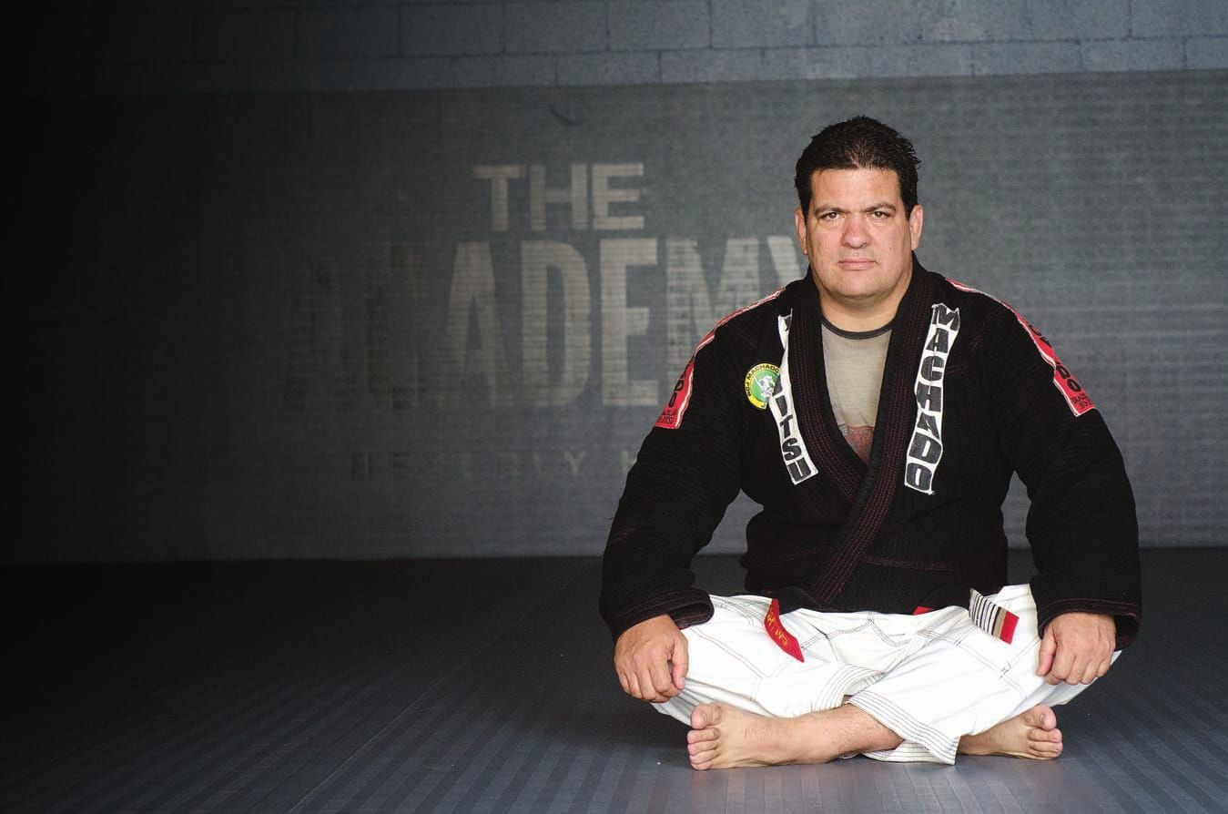 Rigan Machado seated on academy mats in a black gi jacket and white gi trousers.