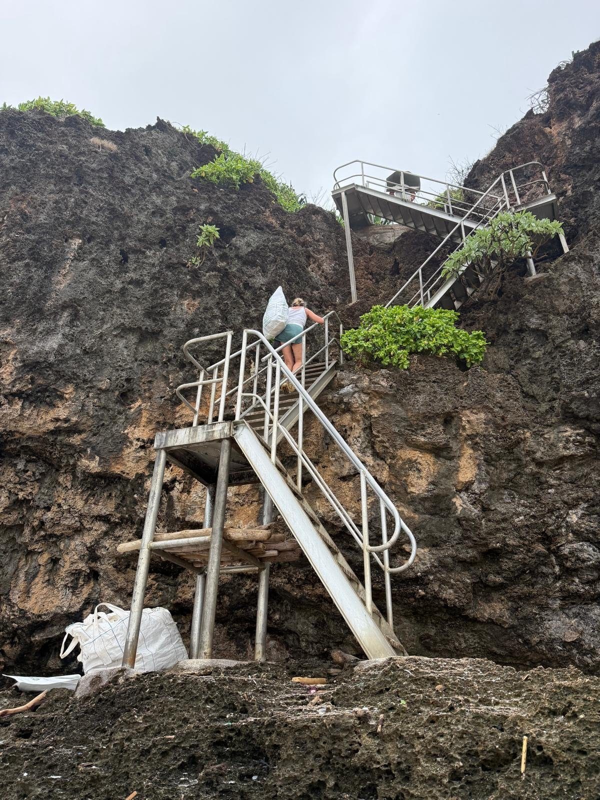 Nicole climbing the steep metal staircase at Greta Beach with a large bag of collected marine debris, photographed by Renee Mouritz