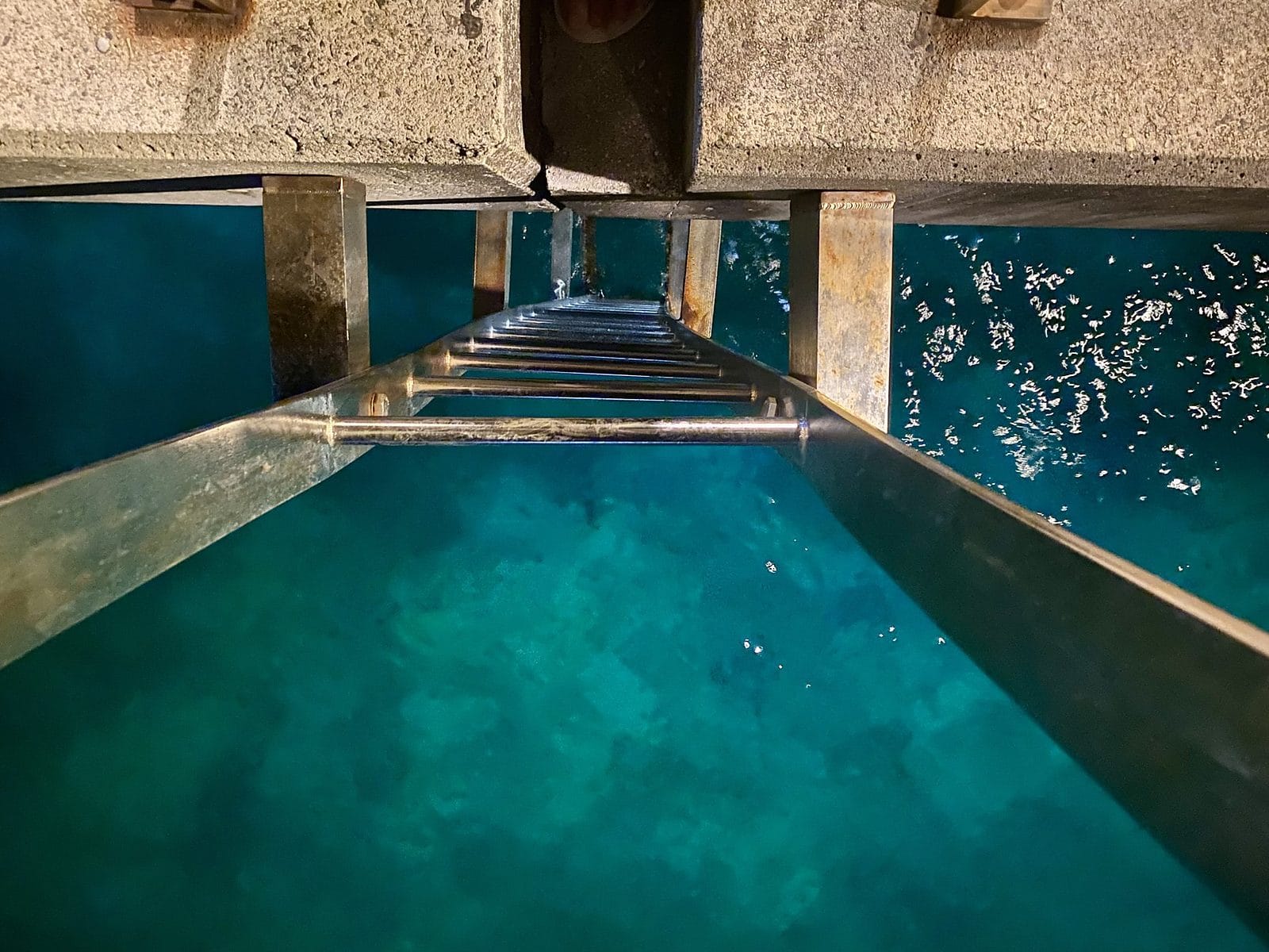 Looking down the metal ladder on the wharf at Flying Fish Cove into clear deep turquoise water, an Earth to Sea News original