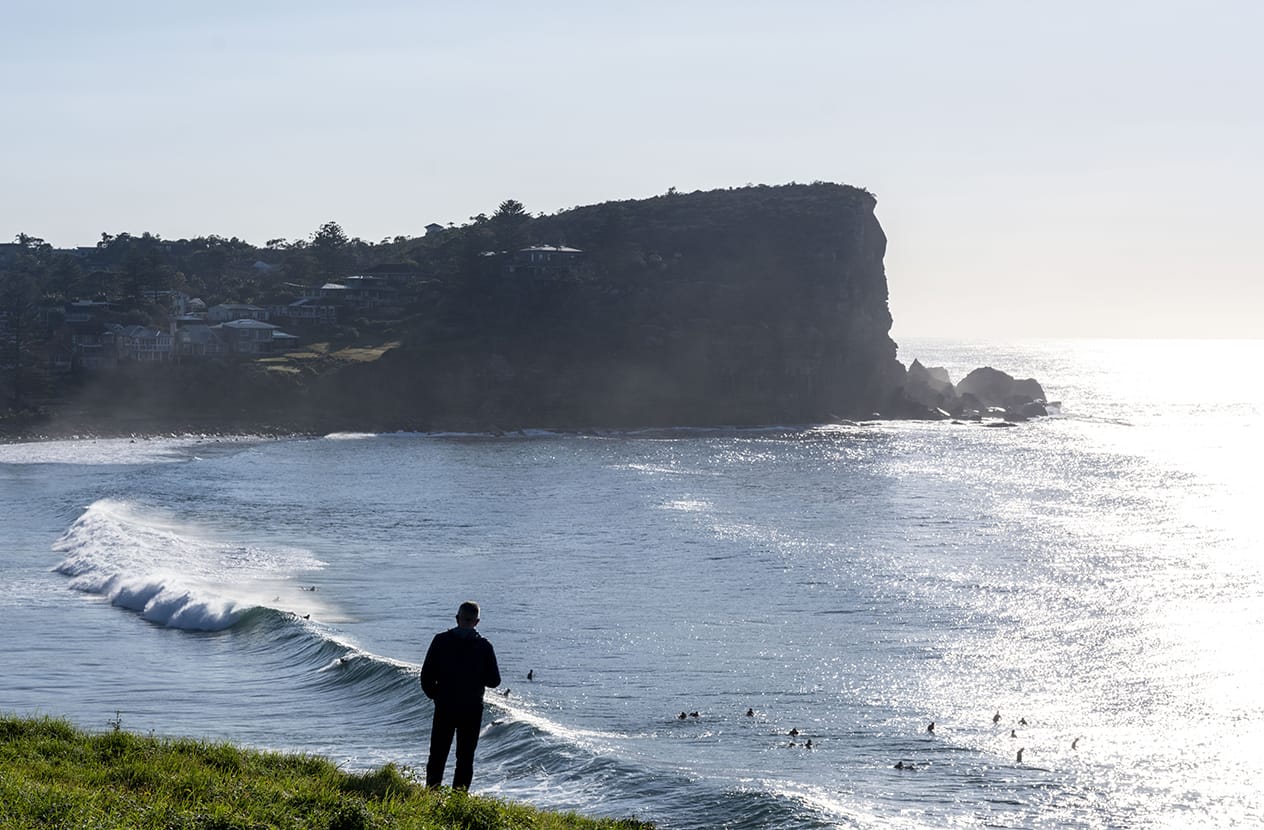 A figure watches the surf from the green headland at Avalon Beach