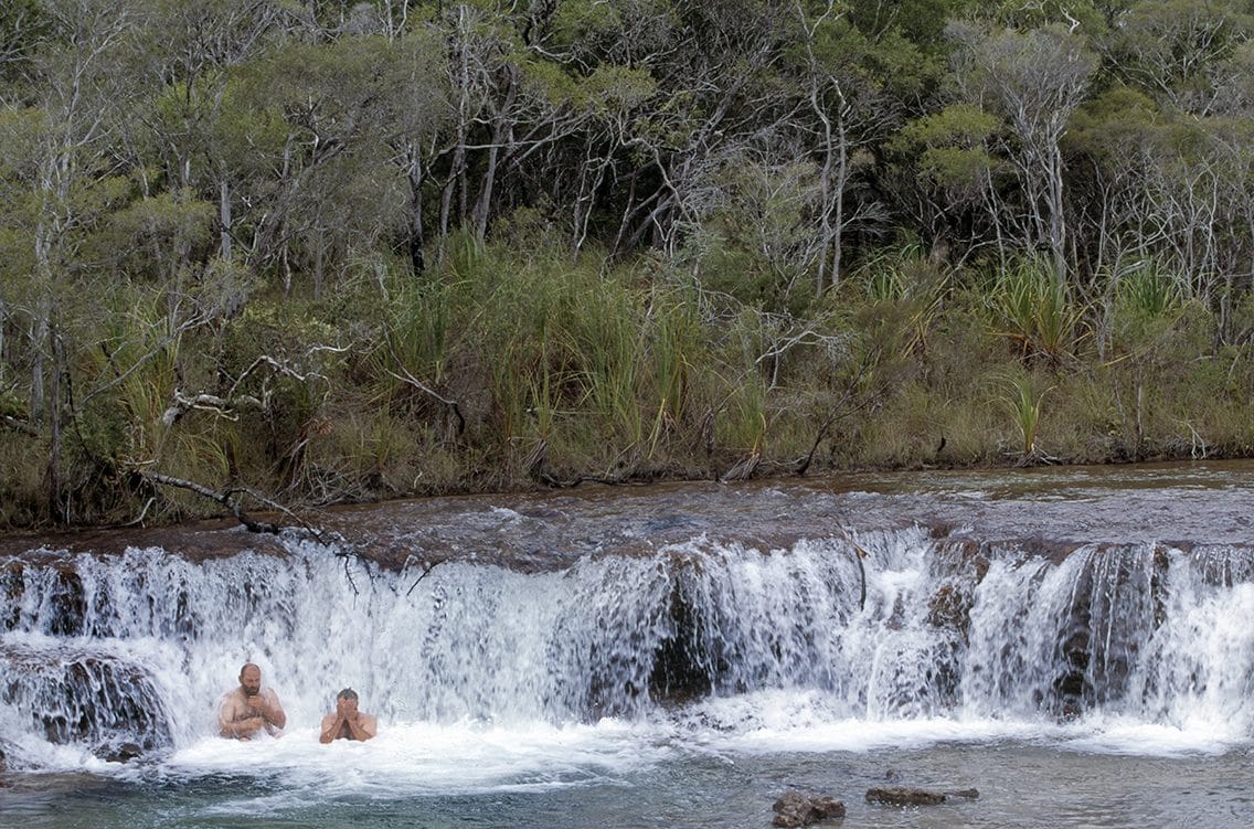 Two swimmers in the pool below a wide bush waterfall on Cape York