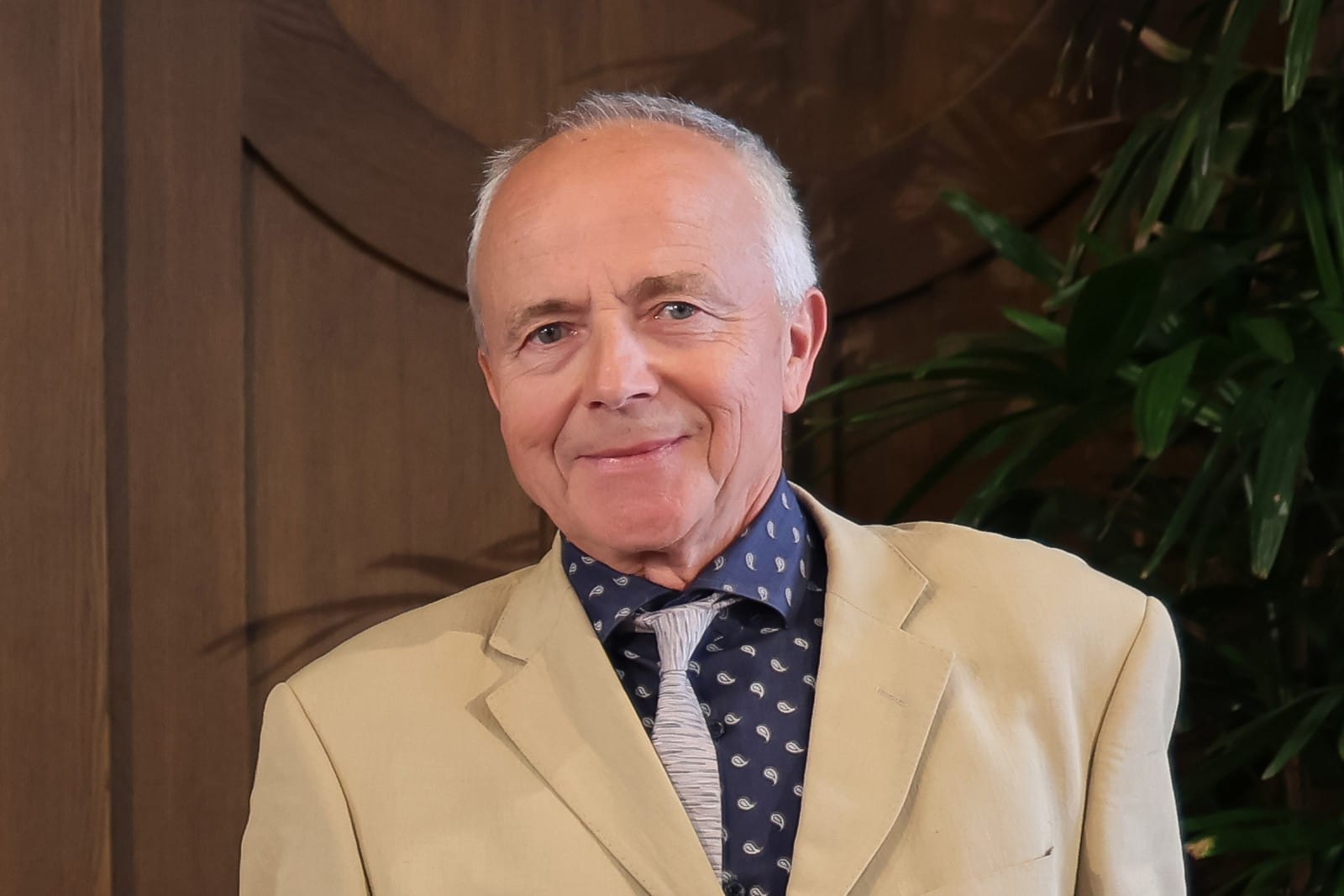 Professor Georg Nagel, smiling, in a cream jacket and patterned blue shirt in front of a wooden wall and a palm