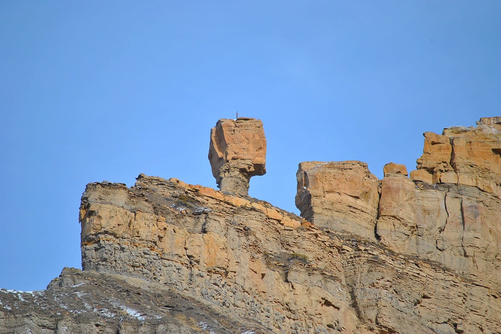 A boulder balanced on a thin pillar of sandstone above a canyon wall under blue sky
